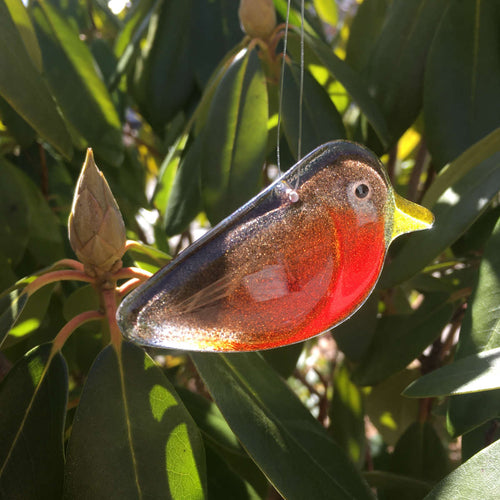 A glossy red and brown glass hanging Robin bird ornament is in the foreground, and set against a background of rhododendron leaves.