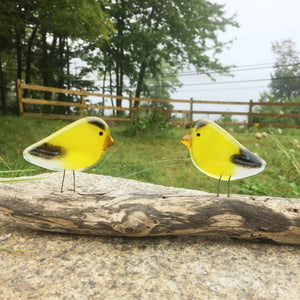two yellow and black glass bird goldfinch ornaments perch on a driftwood log in front of a grassy scene